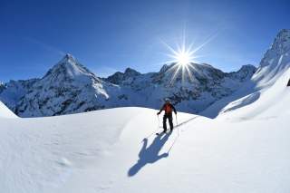 Mensch läuft auf Tourenski durch unberührten Schnee, Sonne steht hinter Gipfeln im Hintergrund