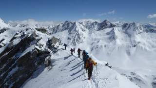 Eine Gruppe von Menschen läuft verschneiten Grat entlang. Im Hintergrund verschneite Bergkette