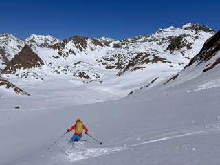 Ein Skifahrer in bunter Ausrüstung fährt eine tief verschneite Berglandschaft mit klar blauem Himmel hinab. Im Hintergrund sind schneebedeckte Gebirgsgipfel zu sehen.