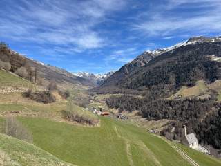 Tal mit grünen Wiesen, kleinen Häusern und einer Kirche, umgeben von bewaldeten Bergen und schneebedeckten Gipfeln unter blauem Himmel.