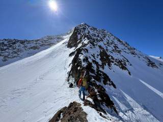 Zwei Bergsteiger mit Skiausrüstung steigen auf einem schmalen, schneebedeckten Grat unter strahlend blauem Himmel auf einen schroffen Gipfel. Die Sonne scheint hell über dem Berg.