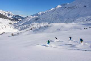 Vier Skifahrer fahren mit hohem Tempo einen flachen Tiefschneehang ins Tal.