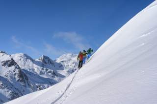 Drei Skitourengeher queren einen steilen Hang vor verschneitem Bergpanorama.