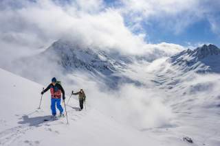 Zwei Skitouengeher im Aufstieg, Berggipfel und Wolken im Hintergrund.