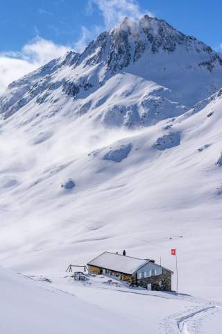 Berghütte aus Naturstein unterhalb eines verschneiten Dreitausenders.