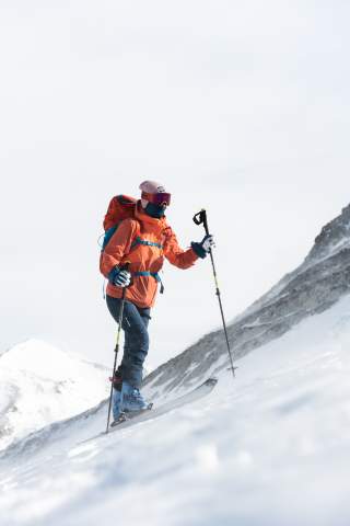 Skitourengeherin von schräg unten im Aufstieg fotografiert. Sie hält die Stöcke in der Hand, trägt eine große Skibrille und hat das Gesicht wegen der Kälte vermummt..