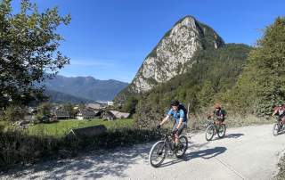 Menschen fahren auf Mountainbikes Schotterstraße entlang, im Hintergrund sieht man ein Dorf