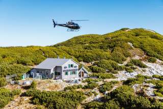 Luftaufnahme einer Alpenvereinshütte zwischen Latschenkiefern, darüber fliegt ein Hubschrauber.