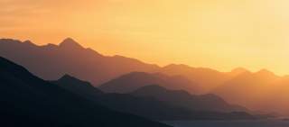 Mehrere Bergsilhouetten in sanften Orangetönen bei Sonnenuntergang.