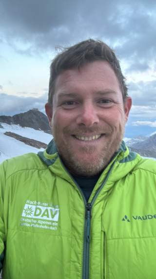Porträt eines lächelnden Mannes mit kurzen braunen Haaren und Bart, bekleidet mit einer grünen DAV- und Vaude-Jacke, vor einer Berglandschaft mit Schnee und bewölktem Himmel.