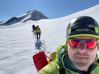 Mehrere Bergsteiger in Ausrüstung sind mit Seilen verbunden und gehen auf einem schneebedeckten Gletscher zu einem Berggipfel, eine Person macht ein Selfie im Vordergrund.
