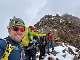 Fünf Bergsteiger in Schutzhelmen und Ausrüstung stehen auf einem schneebedeckten, felsigen Berggipfel, während einer von ihnen ein Selfie macht. Der Himmel ist bewölkt.
