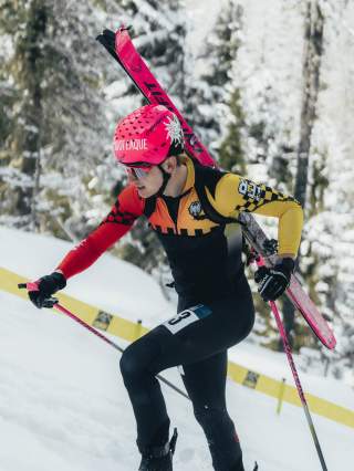 Ein Skimo Athlet mit pinkem Helm und pinken Ski auf dem Rücken läuft im Schnee bergauf. Im Hintergrund Schnee und Wald.