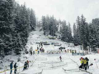 Man sieht den Sprint Parcour, aufgebaut zwischen Bäumen und viel Schnee. Unten sieht man die Diamanten aufgebaut, weiter oben die Tragepassage. Außerdem viele Werbebanner. Darum stehen ein paar Leute, die die Wechsel kontrollieren oder zuschauen.