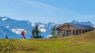 Eine Frau wandert von links über eine Alm auf eine Berghütte zu. Im Hintergrund hohe Alpengipfel.