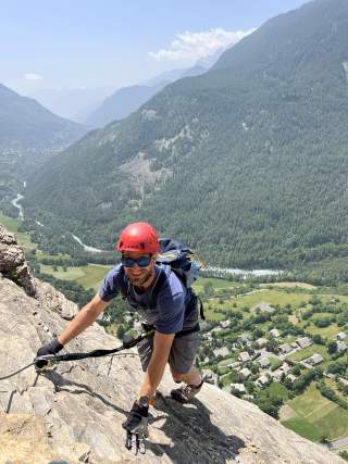 Ein Mann mit rotem Helm und Kletterausrüstung klettert an einer Felswand, im Hintergrund ein Tal mit Fluss, Wäldern und Häusern.