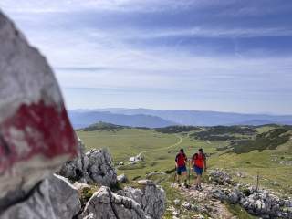 Zwei Wanderergehen auf einem Wanderweg bergauf, etwas unterhalb liegt eine Hütte.