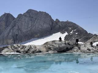 Mehrer Menschen stehen und sitzen am Rand eines Gletschersees, im Hintergrund erheben sich die Berge.