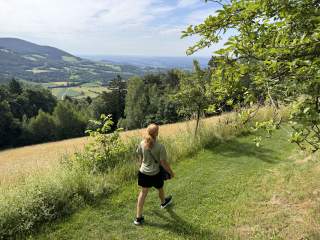 Eine Frau mit Umhängetasche wandert über einen Wiesenstreifen, links der Blick auf Hügelland.