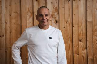 Porträt Volker Schöffl vor Holzwand. Er trägt ein weißes Langarmshirt.