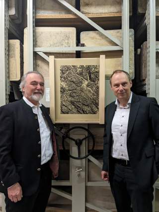 Zwei Männer blicken in die Kamera. Im Hintergrund ist ein Rollregal zu sehen, in dem Lithographiesteine gelagert werden.