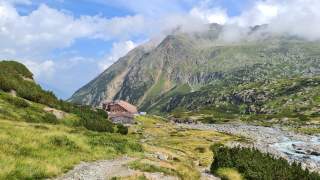 Berglandschaft mit grün bewachsener Wiese, einem kleinen Bach, Wanderweg und zwei Hütten vor steilen Felsen unter leicht bewölktem Himmel.