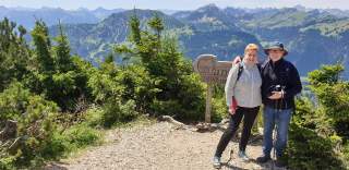 Ehepaar in Bergwandermontur auf einem Gipfel vor Bergpanorama.