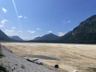 Weitgehend ausgetrockneter Stausee mit einzelnen Booten am kiesigen Ufer, umgeben von bewaldeten Bergen unter blauem Himmel.