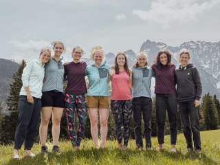 Acht Frauen stehen Arm in Arm auf einer Wiese mit Bergen und Bäumen im Hintergrund und lächeln in die Kamera.