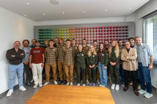 Athletinnen, Athleten und Funktionäre stehen vor einer Wand auf der viele Garnspulen in bunten Farben aufgehängt sind.