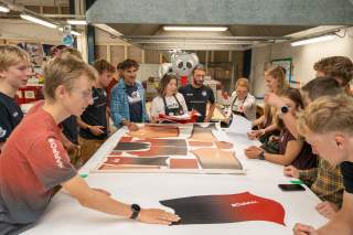 Athletinnen, Athleten und weitere Personen stehen um einen großen Tisch herum. Auf dem Tisch liegt ein Papier mit dem Rennanzug Design.