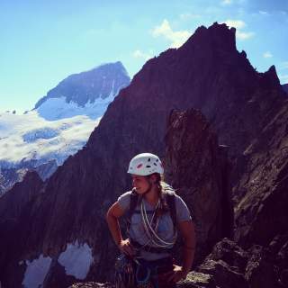 Frau mit Kletterausrüstung und Helm steht auf einem Felsen vor beeindruckenden, schroffen Berggipfeln mit Gletscher im Hintergrund.