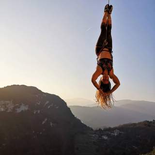 Frau hängt kopfüber und gesichert an einem Seil vor einem Bergpanorama bei klarem Himmel. Die Sonne scheint schwach im Hintergrund und hebt ihre langen Haare hervor.