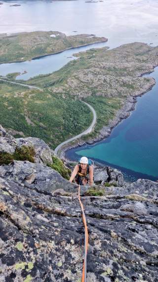 Frau klettert mit Helm und Kletterausrüstung an einem steilen Felsen, im Hintergrund Küste mit grün bewachsenen Klippen und blauem Meer. Ein orangefarbenes Sicherungsseil verläuft vom unteren Bildrand zur Kletterin.