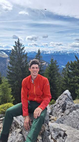Mann in roter Jacke und grüner Hose sitzt auf felsigem Untergrund vor einem Hintergrund aus Tannenbäumen und Bergen mit bewölktem Himmel.