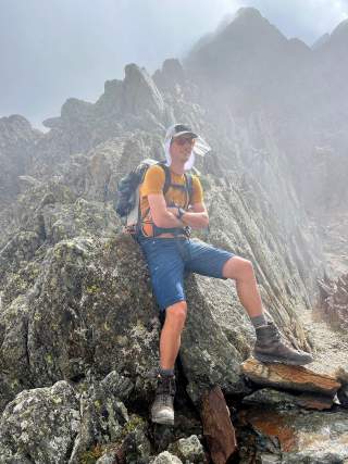 Wanderer mit Rucksack, Sonnenhut und Sonnenbrille sitzt entspannt auf einem großen Felsen in nebliger, felsiger Berglandschaft.