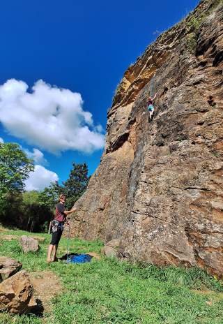 Eine Person klettert an einer Felswand, während unten eine weitere Person die Sicherung mit einem Seil übernimmt, umgeben von grüner Wiese und blauem Himmel mit einigen Wolken.