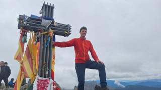 Mann in roter Jacke posiert mit einem großen, mit bunten Tüchern geschmückten Gipfelkreuz auf einem Berggipfel bei bewölktem Himmel.