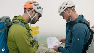 Zwei Männer mit Kletterhelmen stehen in den Bergen und schauen gemeinsam auf ein Kletterstei Topo.