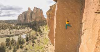 Nina Caprez hängt an einer orangefarbenen Felswand im Smith Rock State Park in Oregon. Im Hintergrund schlängelt sich ein Fluss durch die trockene Landschaft.