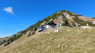 Berglandschaft mit einer Berghütte auf einer Wiese vor einem Hang mit Nadelbäumen, unter klarem blauem Himmel.