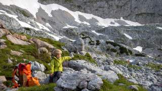 Ein Junge sitzt auf einem Stein in den Bergen und zeigt nach rechts zur Ingolstädter Hütte..
