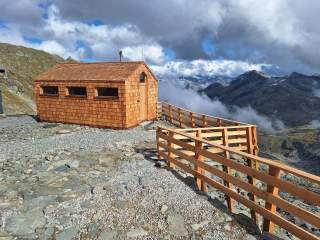 Kleine Berghütte aus Holz mit Schindeldach und drei Fenstern, daneben ein hölzerner Zaun, im Hintergrund Berge und Wolken.