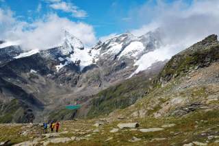 Drei Wanderer stehen auf einem felsigen Bergpfad mit Blick auf schneebedeckte Gipfel und einen kleinen türkisfarbenen See in einem Tal. Der Himmel ist teils bewölkt, teils blau.