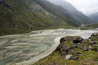 Berglandschaft mit grün bewachsenen Hängen und einem breiten, flachen Flussbett mit mehreren Wasserarmen. Der Himmel ist bewölkt und neblig.