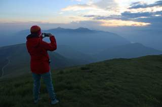 Person in roter Jacke fotografiert mit dem Smartphone eine neblige Berglandschaft bei Sonnenuntergang.