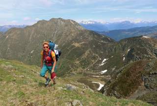 Eine Person mit Rucksack wandert auf einem Bergpfad, im Hintergrund hohe Berge und vereinzelte Schneefelder.