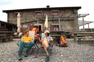 Zwei Frauen sitzen entspannt vor einer Berghütte auf Liegestühlen, stoßen mit Getränken an, neben ihnen stehen große Wanderrucksäcke.