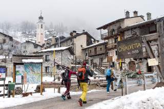 Drei Skifahrer mit Ausrüstung gehen durch das verschneite Dorf Chialvetta, umgeben von traditionellen Steinhäusern und einer Kirchturmspitze im Hintergrund. Ein hölzernes Schild mit der Aufschrift „Chialvetta m. 1495“ steht an einem Holzbalken am Wegrand.