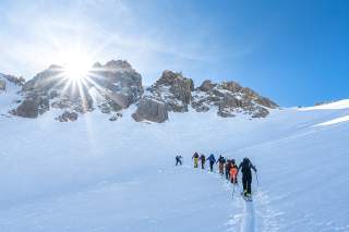 Gruppe von Skitourengehern steigt bei sonnigem Wetter eine verschneite Bergflanke unter felsigen Gipfeln empor.
